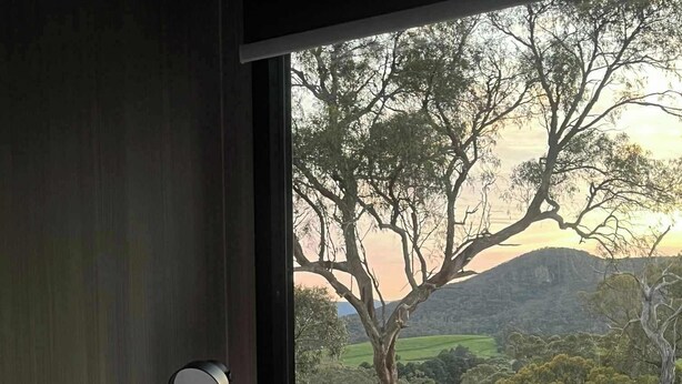 Een slaapkamer met een bed, een bedlampje en uitzicht op een landelijk landschap met bomen en heuvels.