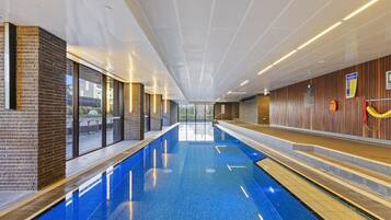 A modern indoor swimming pool with clear glass walls, wooden paneling, and a bright ceiling.