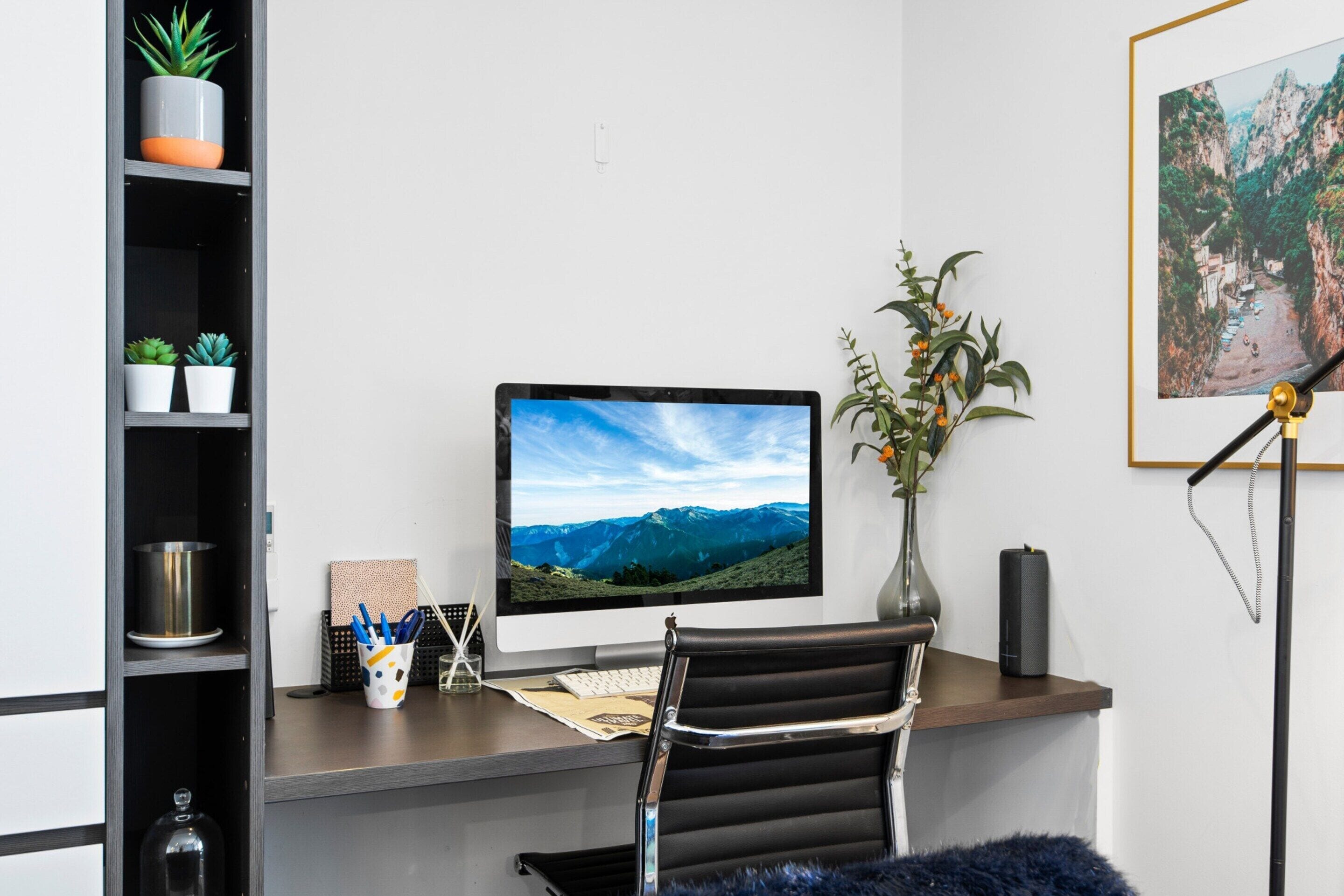 A modern office desk with a computer monitor, a vase with flowers, and a framed picture on the wall.