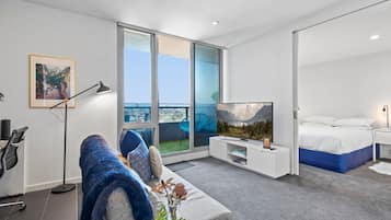 A modern hotel room with a large window, a bed, a sofa, a TV, and a desk with a lamp.