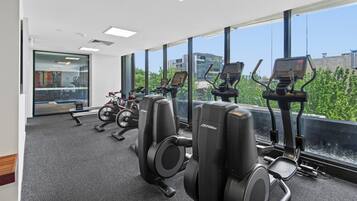 A gym with cardio machines, a weight bench, and large windows offering a city view.