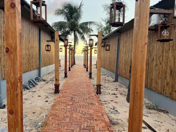 Ein Weg führt zu einem Strandbereich mit Bambuswänden und hängenden Laternen.