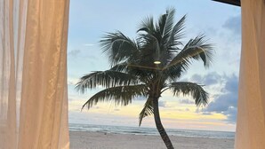 A room with a wooden table and chairs, a window with a view of the beach and a palm tree, and a wooden fence outside.