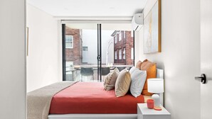 A modern bedroom with a large window, a bed with red bedding, a bedside table with a lamp, and a view of the outside. - Manly Muse with Water View and Lock Up Garage (Manly)