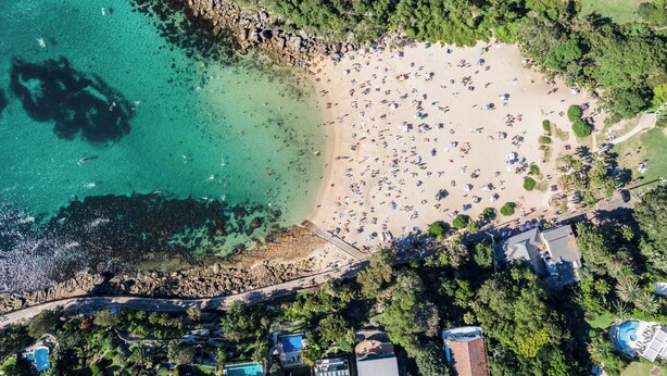 An aerial view of a beach with clear turquoise waters, a sandy shore, and numerous umbrellas. There are buildings with pools and a dense tree line in the background. - Manly Muse with Water View and Lock Up Garage (Manly)