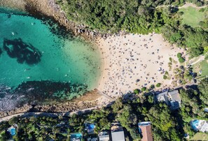An aerial view of a beach with clear turquoise waters, a sandy shore, and numerous umbrellas. There are buildings with pools and a dense tree line in the background. - Manly Muse with Water View and Lock Up Garage (Manly)