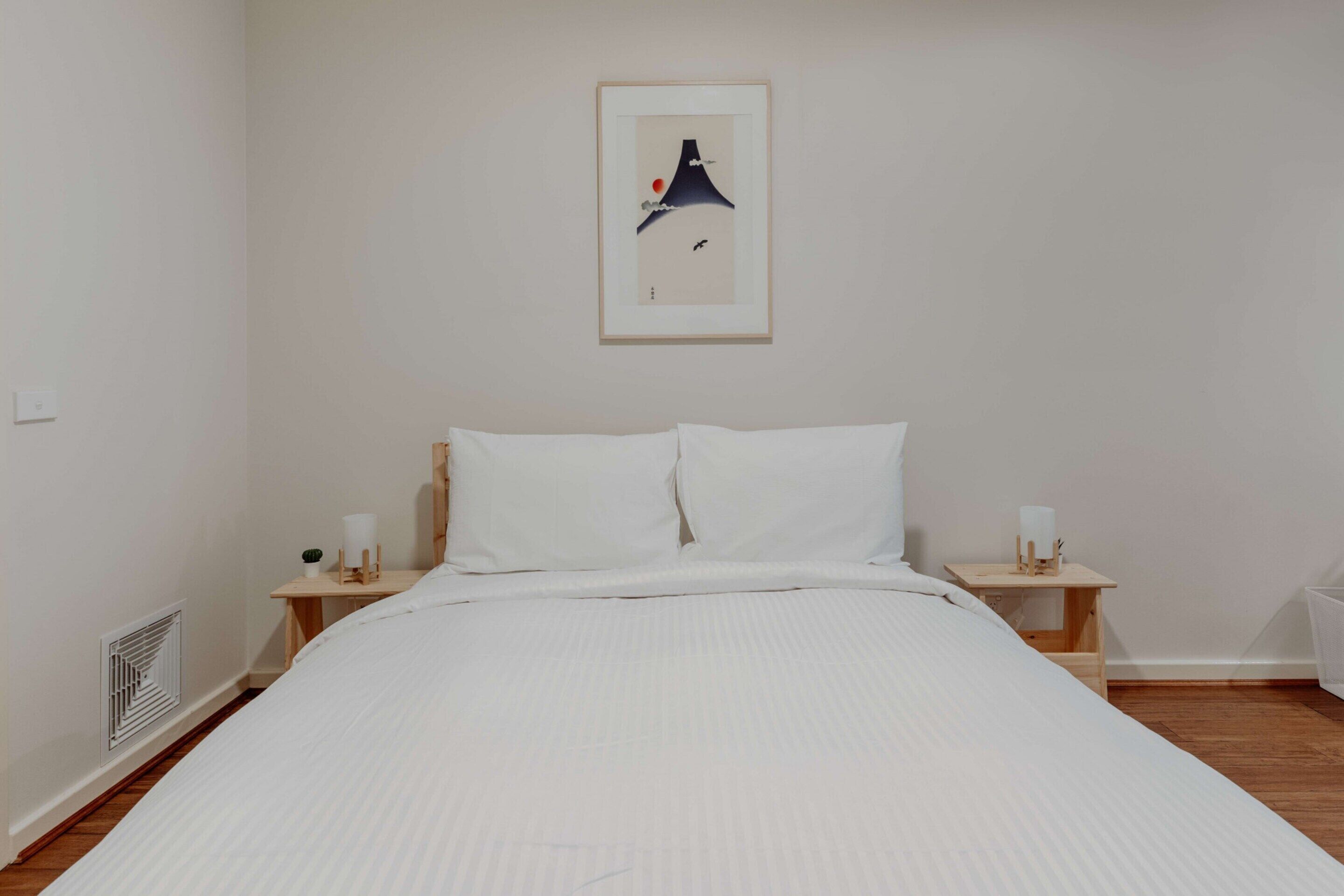Una cama bien hecha con ropa de cama blanca, una obra de arte enmarcada encima y una mesita de noche de madera pequeña con una lámpara a cada lado.