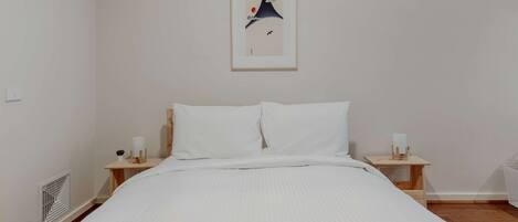 A neatly made bed with white linens, a framed artwork above it, and a small wooden nightstand with a lamp on each side.