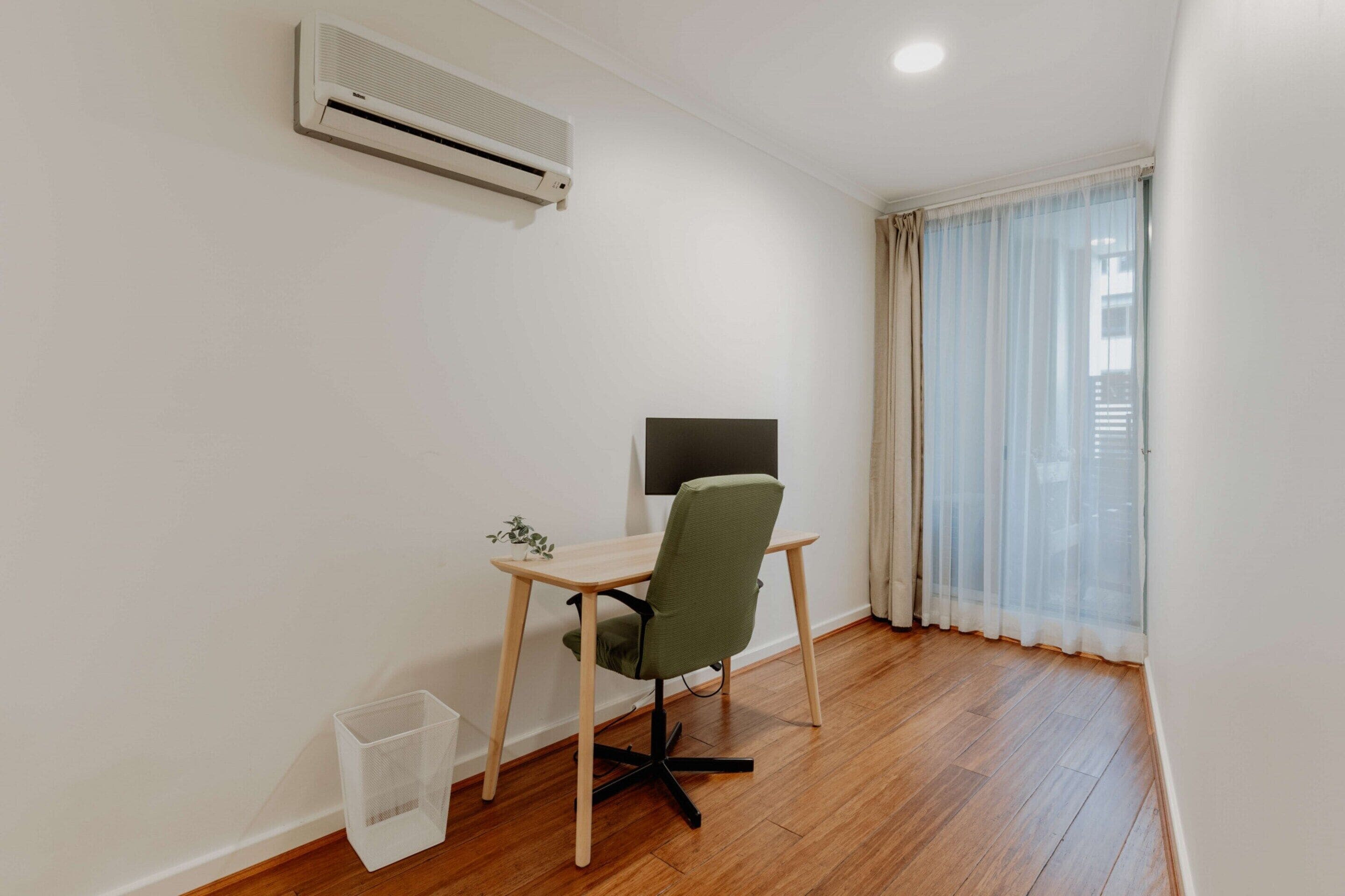 Una habitación pequeña y limpia, con piso de madera, un escritorio con silla, un televisor y una ventana con cortinas traslúcidas.
