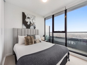 A modern bedroom with a large bed, a city view through floor-to-ceiling windows, and a minimalist design.