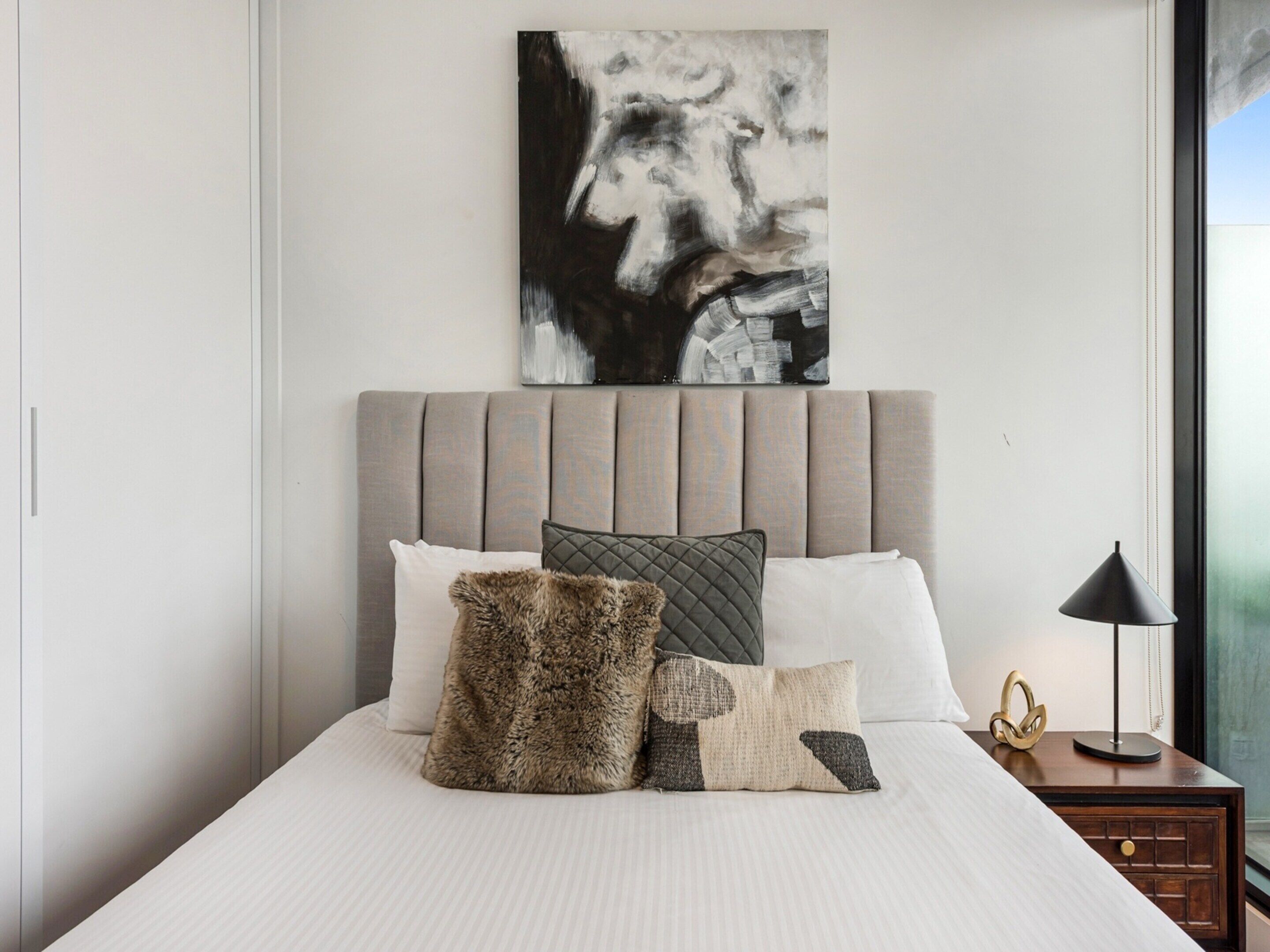 A neatly made bed with a grey upholstered headboard, a variety of pillows, and a modern abstract painting above it.