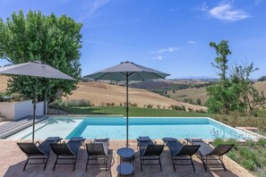 A swimming pool with lounge chairs and umbrellas, surrounded by a hilly landscape.