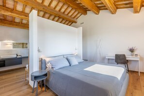 A bedroom with a large bed, a bedside table, a chair, a desk, and a bathroom with a sink and mirror.