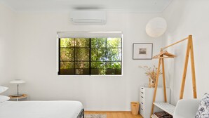 A modern bedroom with a large window, a bed, a bedside table with a lamp, a chair, and a small side table with decorative items. - Spacious 2-bed Townhouse With a Lush Courtyard (Sydney)