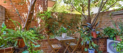 A patio with a brick wall, potted plants, a wooden table, and chairs.