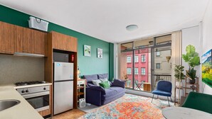Une cuisine compacte équipée d’un réfrigérateur, d’une cuisinière et d’un four. Un canapé et une table à manger sont placés près d’une grande fenêtre qui offre une vue sur un bâtiment rouge.