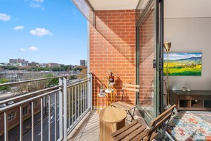 A balcony with a wooden table and chairs, a brick wall, and a painting of a landscape. - Chic Inner-city 1 Bedroom With Balcony (Sydney)