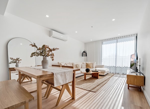 A modern living room with a wooden dining table, sofa, and a large window with sheer curtains.