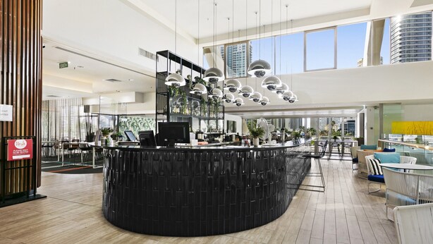 A modern hotel lobby with a central black reception desk, multiple seating areas, and large windows offering city views. - Ocean View 2-bed Studio In the Heart of Surfers (Gold Coast)