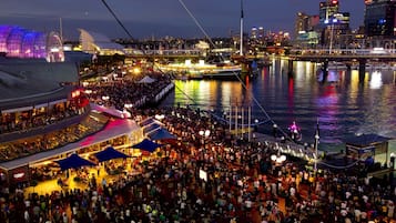 Un evento al aire libre concurrido, con un escenario y carpas, con vistas a una masa de agua, un puente y el horizonte de la ciudad de fondo.