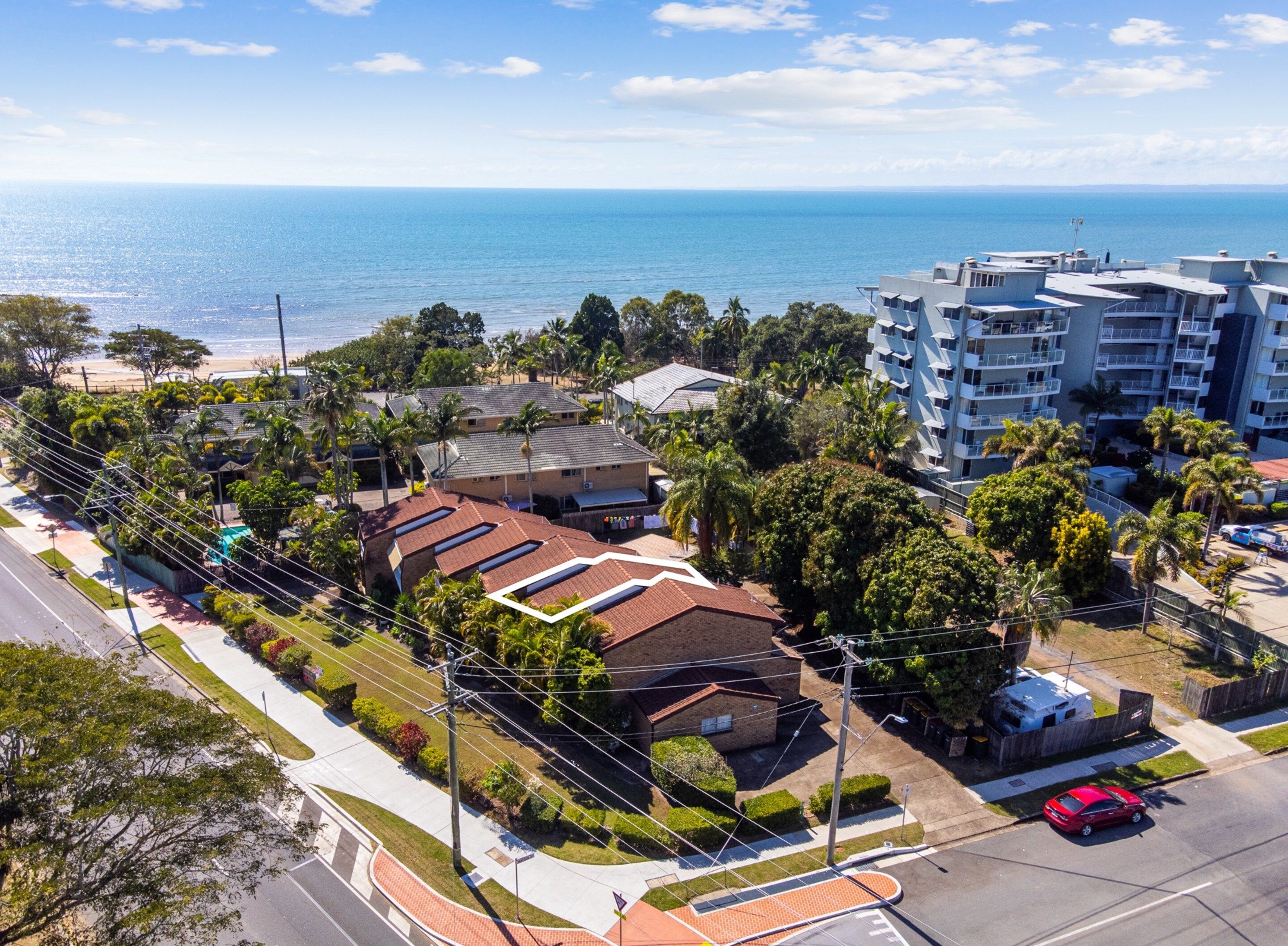 A coastal residential area with a mix of single and multi-story buildings, a road with a red car, and a view of the ocean.