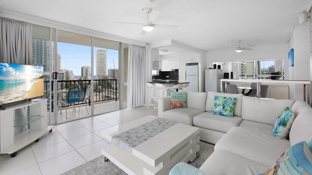 Ein modernes Wohnzimmer mit einem weißen Sofa, einem Couchtisch und einem Fernseher. Ein Balkon bietet Blick auf die Stadt und die umliegenden Gebäude.