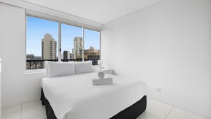 Ein modernes Hotelzimmer mit einem großen Bett, weißer Bettwäsche, Blick auf die Stadt durch ein großes Fenster und einem Balkon mit Geländer.