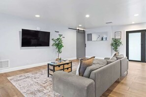 Ein modernes Wohnzimmer mit einem grauen Ecksofa, einem flachbildfernseher an der Wand, einem kleinen Beistelltisch mit Büchern und einer Pflanze sowie einem großen Fenster mit Ausblick.