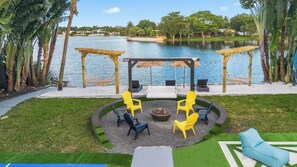 An outdoor seating area with a fire pit, surrounded by chairs, overlooking a lake with a gazebo and palm trees.