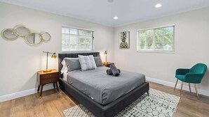 Ein Schlafzimmer mit einem großen Bett, zwei Nachttischen, einem Beistelltisch, einem blaugrünen Sessel und einem Fenster mit Jalousien.