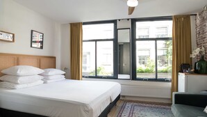 A hotel room with a large bed, a window with curtains, a radiator, a small table with a vase, and a green armchair.