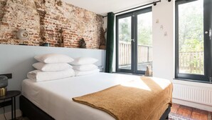 A bedroom with a brick wall, a large bed with white linens, a brown throw blanket, and a view of a balcony through a sliding door. - Twenty Five Garden (Room 2) (Amsterdam)