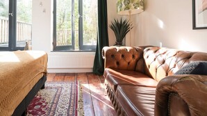 A room with a bed, a brown leather sofa, a patterned rug, a floor lamp, and a window with a view of greenery. - Twenty Five Garden (Room 2) (Amsterdam)