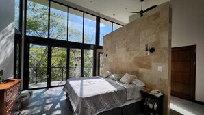 A modern bedroom with a large bed, a dresser, a nightstand, and a view of trees through large windows.