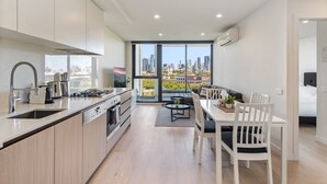 A modern kitchen with a large island, a dining area with a table and chairs, and a view of the city skyline through large windows. - City Views & Inner-north Cool at Your Door (Melbourne)