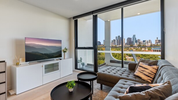 A modern living room with a large TV, a sofa, a coffee table, and a view of a city skyline through large windows. - City Views & Inner-north Cool at Your Door (Melbourne)