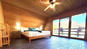 A bedroom with a wooden ceiling, a bed with pillows, a nightstand, and a large window. - Family Cabin w/Hot Tub | Red Rock Views | Pets (Kanab)