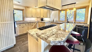 A kitchen with wooden cabinets, a granite countertop, a central island with bar stools, and large windows. - Family Cabin w/Hot Tub | Red Rock Views | Pets (Kanab)