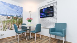 A modern room with a white brick wall, a mounted TV, a small table with chairs, and a view of the beach through a large window. - Gemuetliche Ferienwohnung mit Wintergarten, Bensersiel (Esens)
