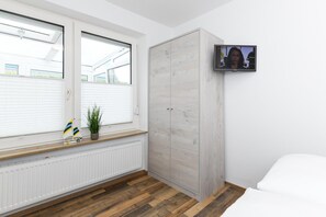 Ett sovrum med en trägarderob, en tv som är monterad på väggen, ett fönster med persienner, ett element och en liten växt på fönsterbrädan.