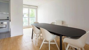 Un comedor moderno con una mesa negra y sillas blancas, una cocina con electrodomésticos de acero inoxidable y un amplio ventanal con vistas a zonas verdes.