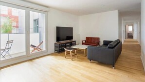 Un amplio salón con un ventanal grande, un televisor de pantalla plana, un sofá, una mesa de centro y un taburete pequeño.
