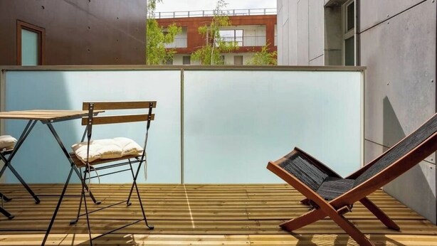 Una terraza de madera con una mesa y sillas, una silla lounge y vistas a un edificio con balcones y vegetación.