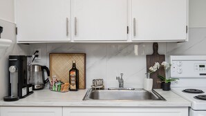 Una cocina moderna con armarios blancos, un lavamanos de acero inoxidable, una cafetera, una botella de aceite de oliva y una orquídea en maceta.