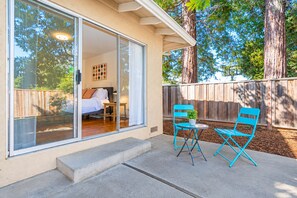 A house with a sliding glass door leading to a bedroom with a bed and a nightstand. There is a small patio with two blue chairs and a table. - Central 3BR Home - Steps from Mountain View DT, Castro St (Mountain View)