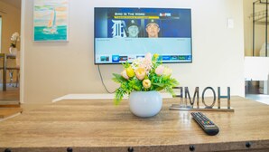 A wooden table with a vase of flowers, a remote control, and a TV displaying a sports channel. - Central 3BR Home - Steps from Mountain View DT, Castro St (Mountain View)