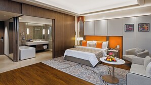 A modern hotel room with a large bed, a sitting area, a small table with fruit, a bathroom with a shower, and a large mirror. - Fortune Select Siliguri - Member ITC Hotels' Group (Jalpaiguri)
