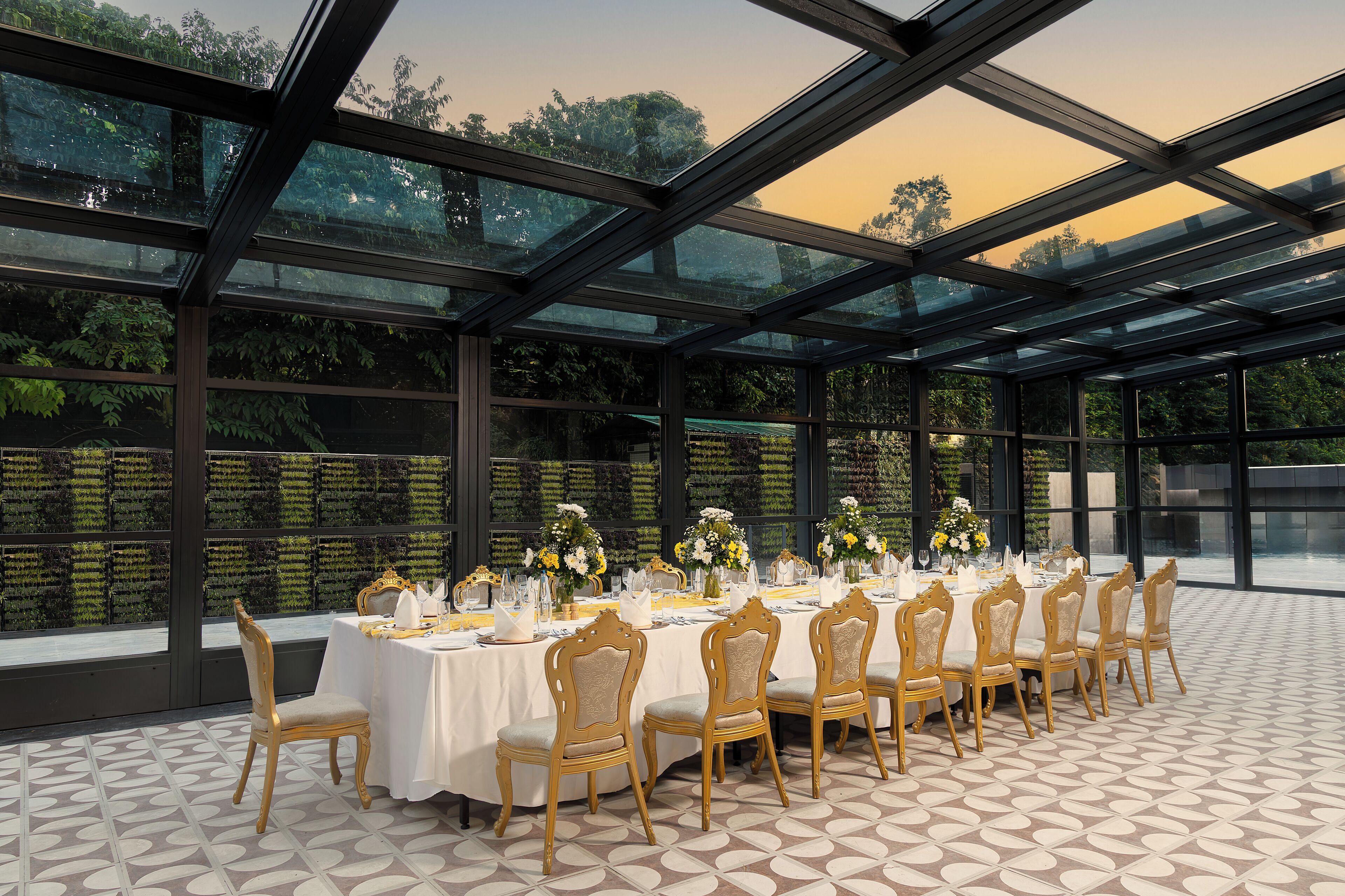 Una larga mesa dispuesta para un evento formal, con sillas, techo de cristal y vistas a la vegetación.