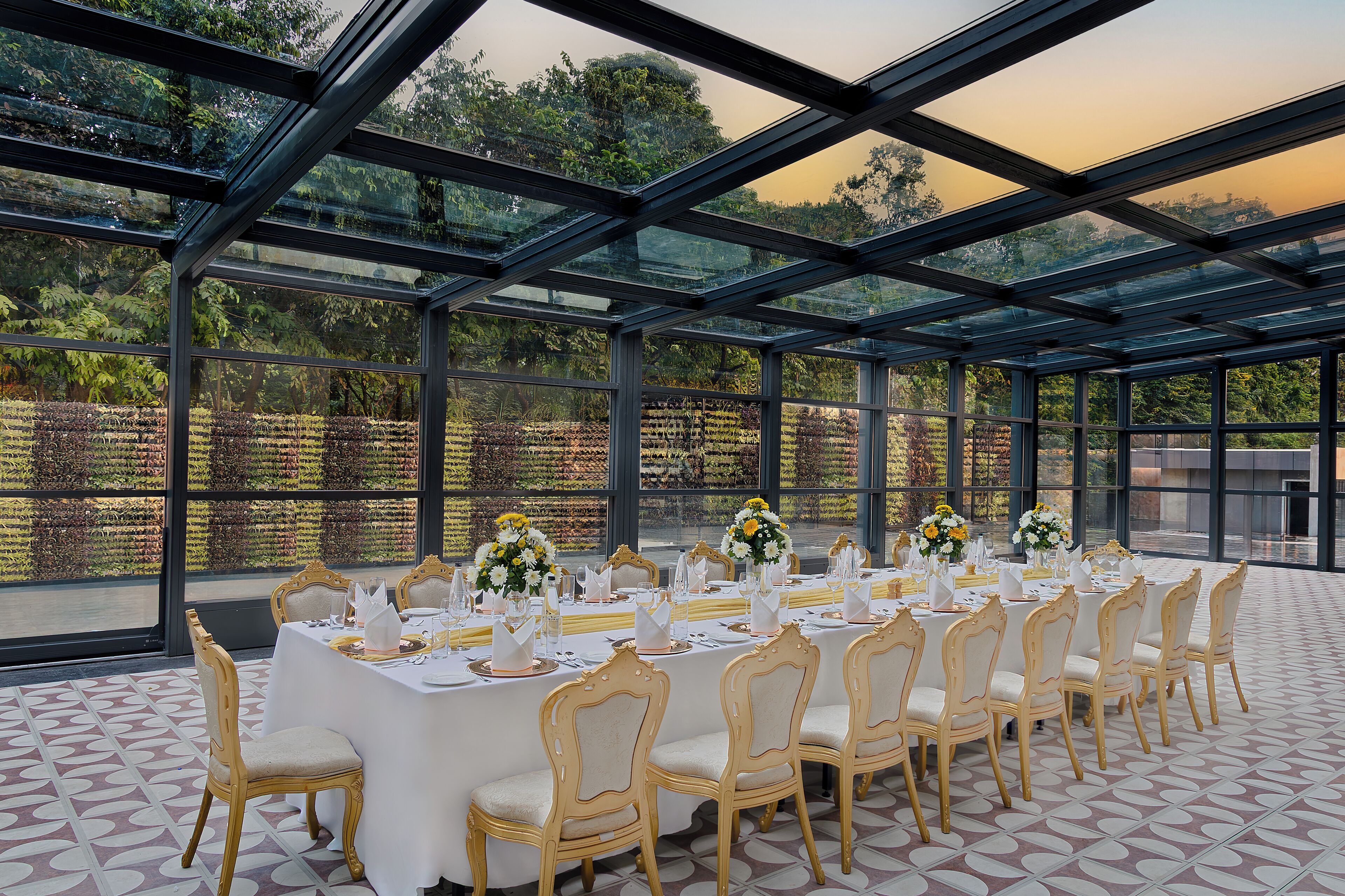 Una larga mesa dispuesta para un evento formal, con sillas, techo de cristal y vistas a zonas verdes.