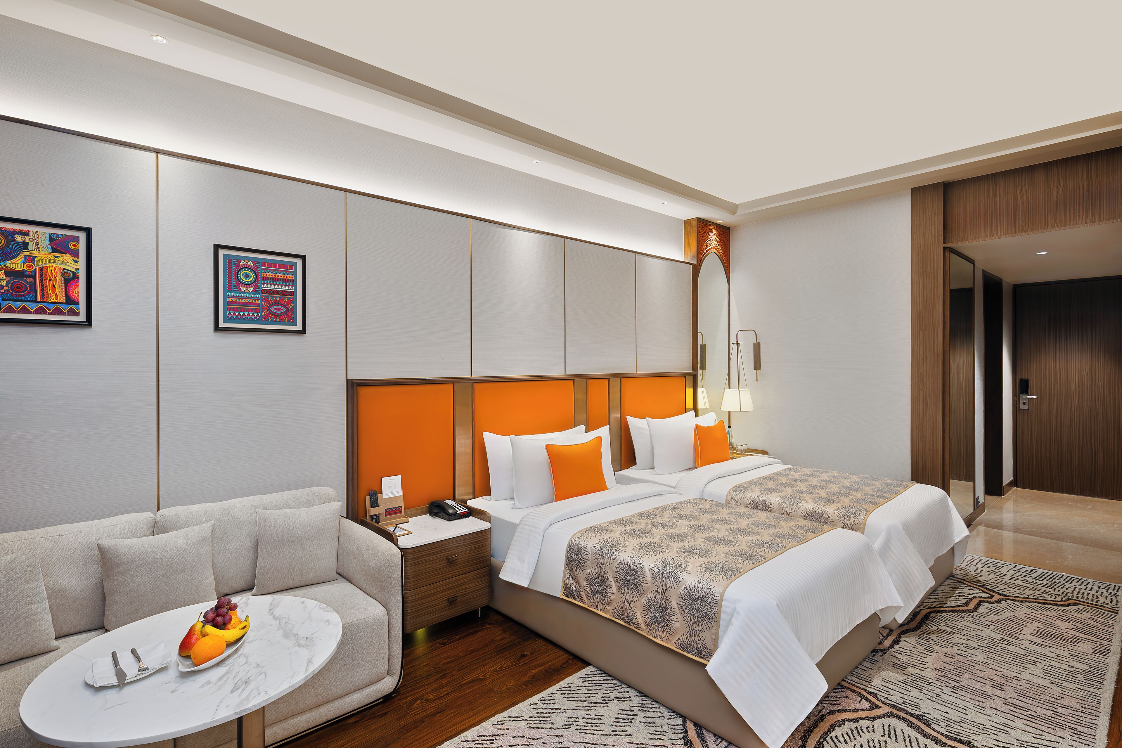 A hotel room with a sofa, two beds, a small round table with fruit, and artwork on the walls.